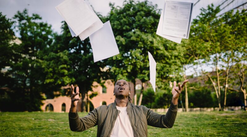 Overjoyed African American graduate tossing copies of resumes in air after learning news about successfully getting job while sitting in green park with laptop