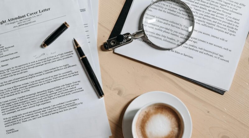 A top view of coffee and job application documents with a magnifying glass on a wooden desk.